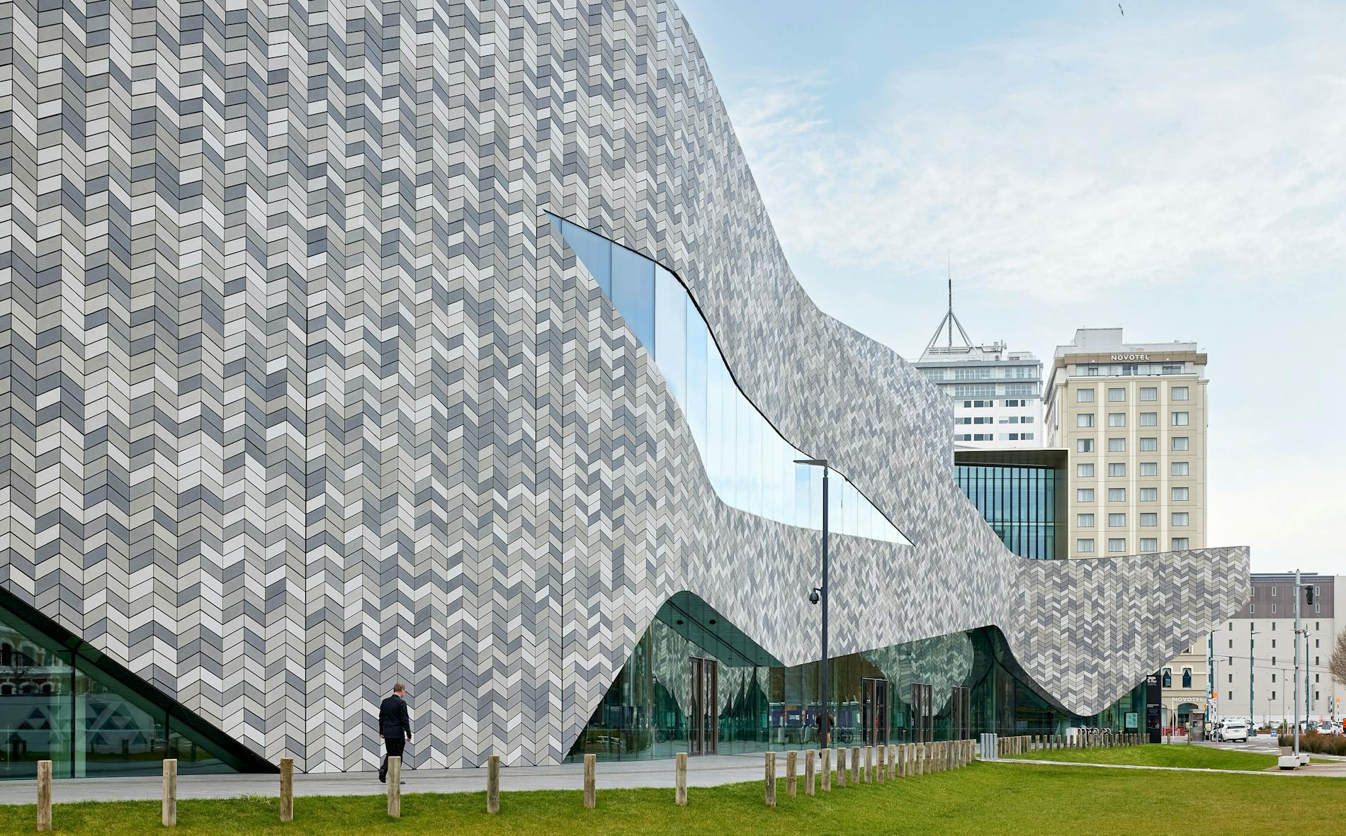 Te Pae, a modern architectural structure with a textured facade, stands prominently as a man walks past, surrounded by a landscaped area and urban buildings.